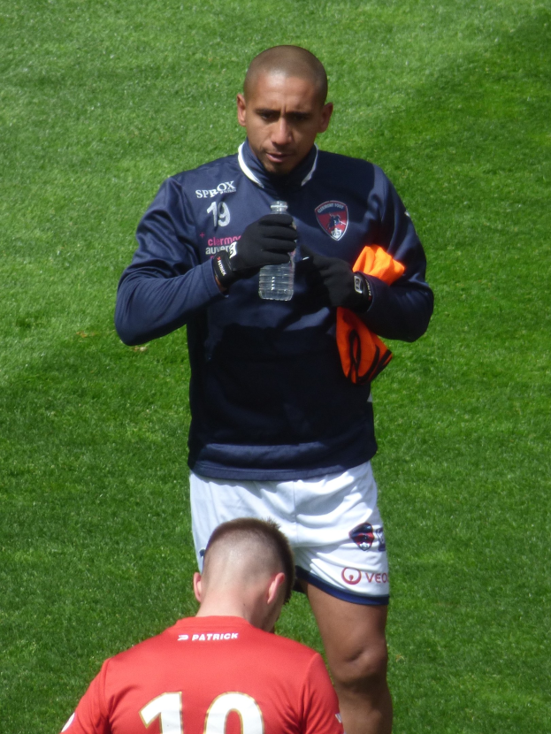 Ein professioneller Athlet steht auf einem grünen Feld und hält eine Flasche in einer Hand und ein oranges Tuch in der anderen.