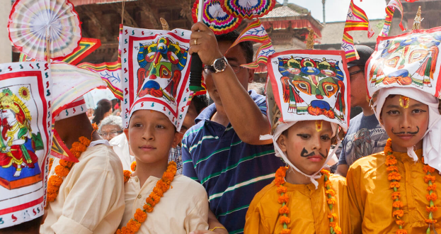 Eine Gruppe von Kindern in bunten Kostümen und Kränzen steht zusammen und hält Fahnen und Gegenstände, mit Gebäuden und Polen im Hintergrund unter einem klaren blauen Himmel, während eines Festivals in Kathmandu.