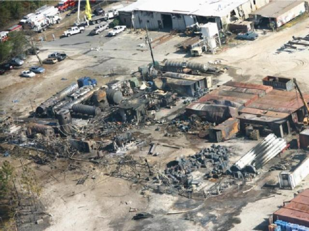 Luftaufnahme eines großen Industriegebiets mit umfangreichen Trümmern, darunter Gebäude, Schuppen, Fahrzeuge, Masten und Bäume, das sich um eine Raffinerie auf einem riesigen offenen Gelände erstreckt.