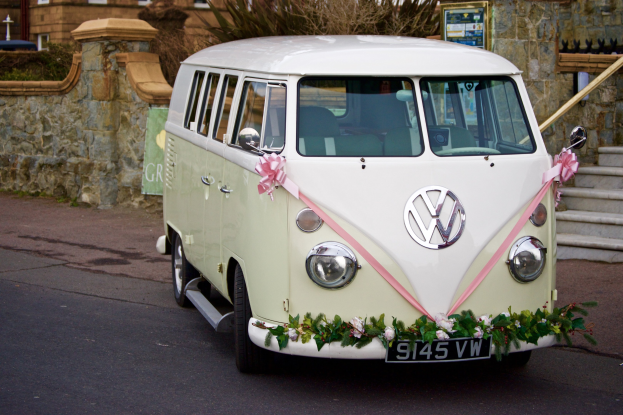 Ein weißer und pinkfarbener VW Camper Van mit floralen Verzierungen, der vor einer Steinwand geparkt ist, neben einer Treppe und einem Gebäude.