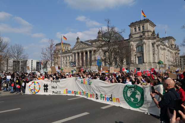 Eine große Gruppe von Menschen geht eine Straße in Berlin, Deutschland, entlang und hält eine Fahne und Flaggen hoch; eine Person hält ein Mobiltelefon; die Straße ist von Bäumen gesäumt und von Gebäuden unter einem bewölkten Himmel umgeben.