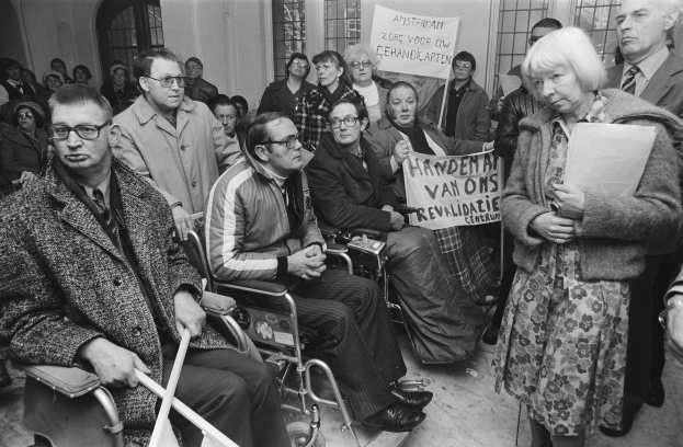 Eine Gruppe von Menschen in Rollstühlen und Stehenden, einige mit Schildern, vor einer Menge, mit Fenstern und einer Wand im Hintergrund, in Schwarz-Weiß abgebildet.