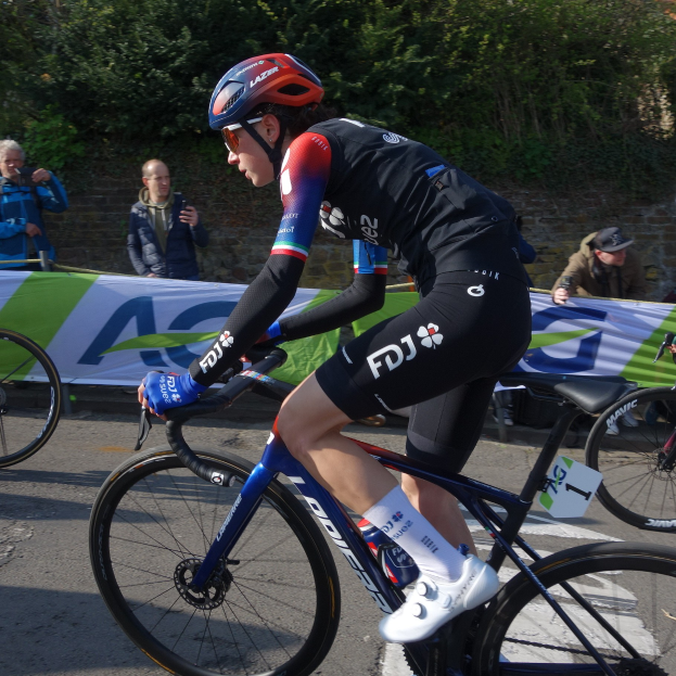 Professionelle Radrennfahrerin Katie Archibald trägt Helm, Schutzbrille, Handschuhe und Schuhe und fährt ein Fahrrad auf einer Straße neben einer Menschenmenge, mit einem Banner, einer Wand, Bäumen und einem Gebäude im Hintergrund.