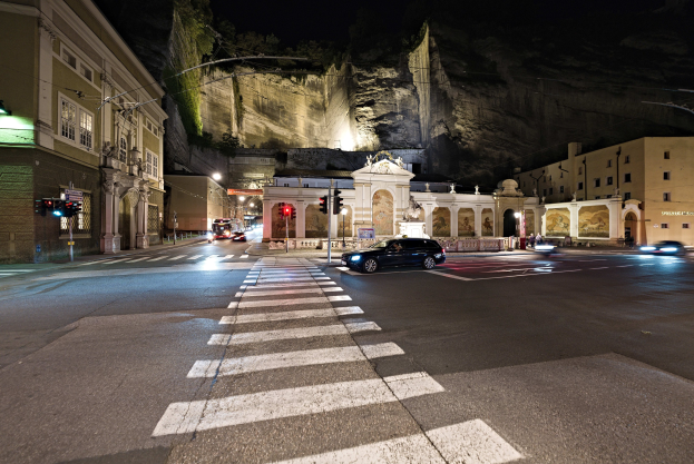 Eine nächtliche Stadtstraße mit fahrenden Autos, Straßenlaternen, Ampeln, beleuchteten Fenstern in Gebäuden, Laternenpfählen, einem Hügel im Hintergrund und einer Zebrastreifenkreuzung unten.