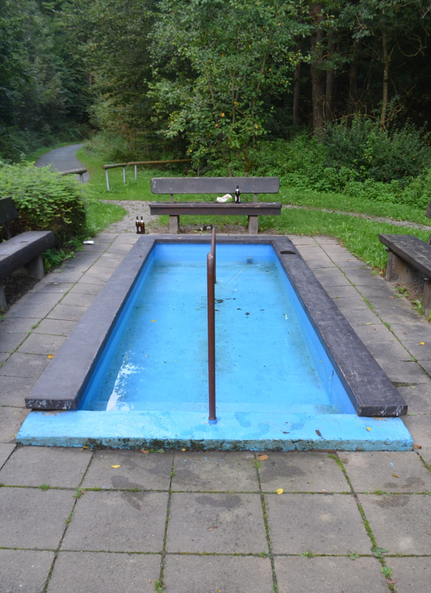 Kleiner Parkpool mit zentralem Pfahl, umgeben von B├Ąnken, Pflanzen, Rasen, Stra├če und B├Ąumen.