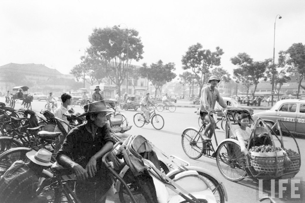 Eine vielbefahrene Straße mit Fahrrädern, Autos und anderen Fahrzeugen, die vorbeifahren, mit Bäumen und Gebäuden im Hintergrund.