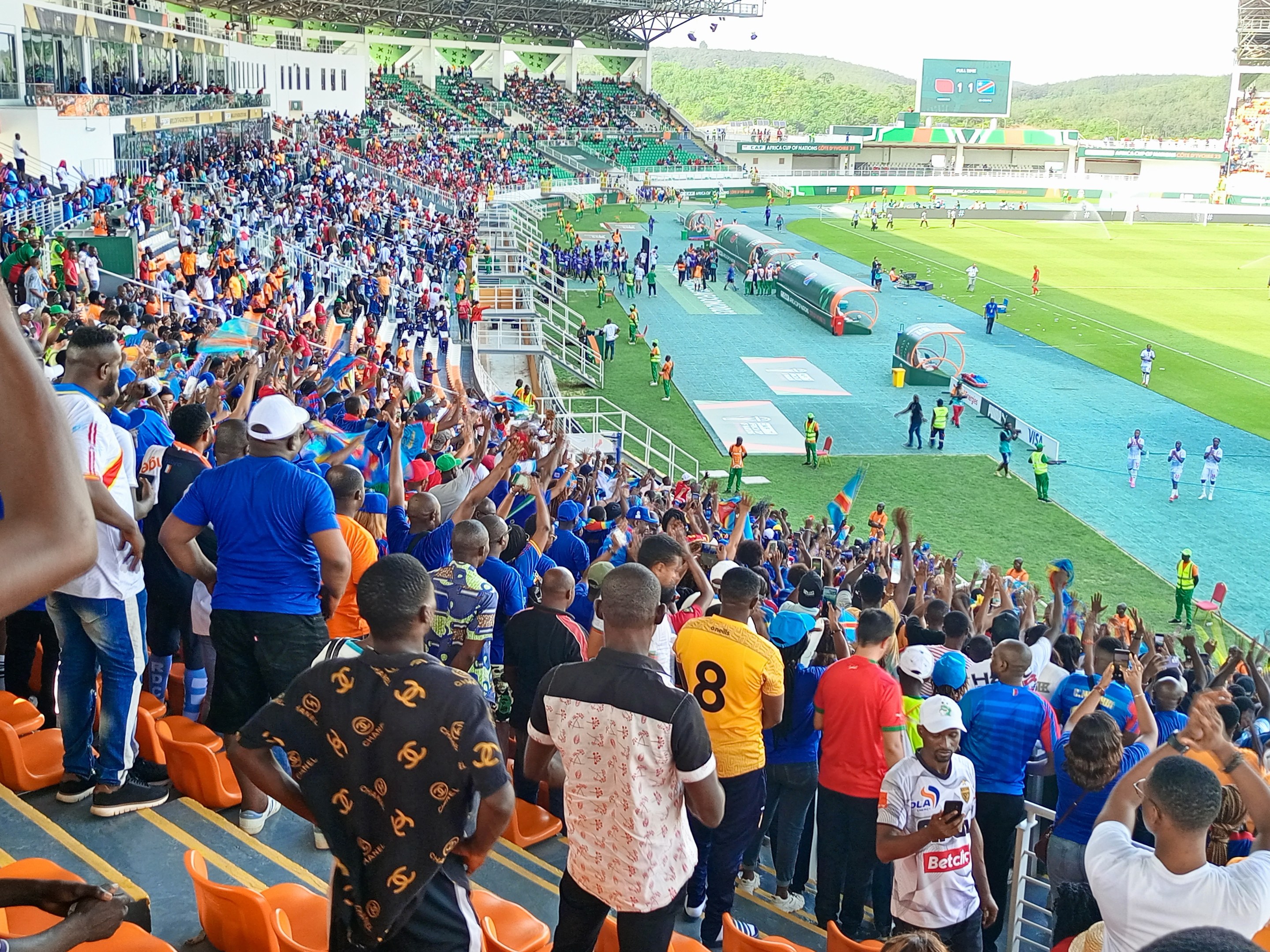 Eine große Menschenmenge versammelt sich vor einem eingezäunten Fußballfeld, wobei einige auf Stühlen sitzen und andere stehen, vor einem Hintergrund von Hügeln und einem klaren blauen Himmel.