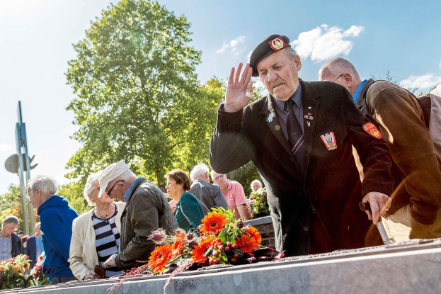 Eine Gruppe von Menschen steht um ein Denkmal herum, ein Mann in militärischer Uniform salutiert an einem Grab, umgeben von Blumensträußen, Bäumen und einem klaren blauen Himmel.