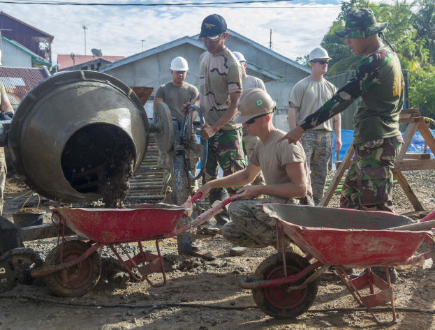 Gruppe von Männern mit Helmen auf einer Baustelle, einige schieben Karren und einer bedient einen Betonmischer, mit Gebäuden, Bäumen, Strommasten und einem bewölkten Himmel im Hintergrund.