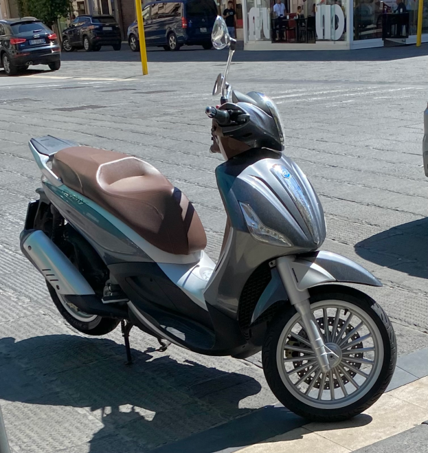 Ein Motorroller steht am Straßenrand mit einem Helm darauf, mit Gebäuden, Bäumen, Polen, Menschen, Stühlen, Tischen und anderen Gegenständen im Hintergrund.