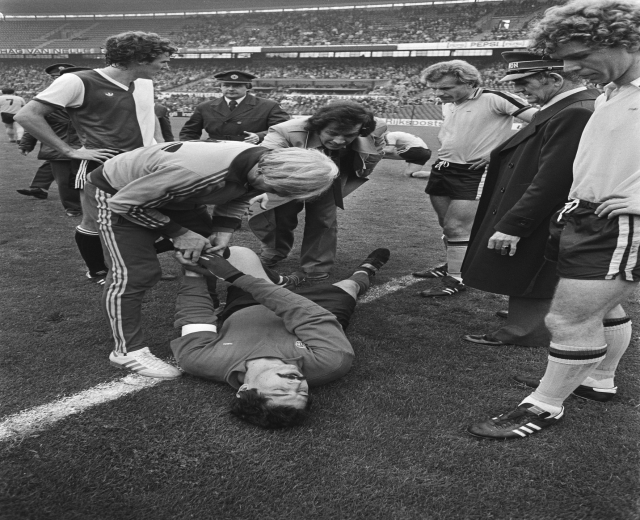 Schwarzes und weißes Foto eines Fußballspielers, der auf dem Boden liegt und von einer Gruppe Männer umgeben ist, mit einer Menge im Hintergrund eines Stadions.