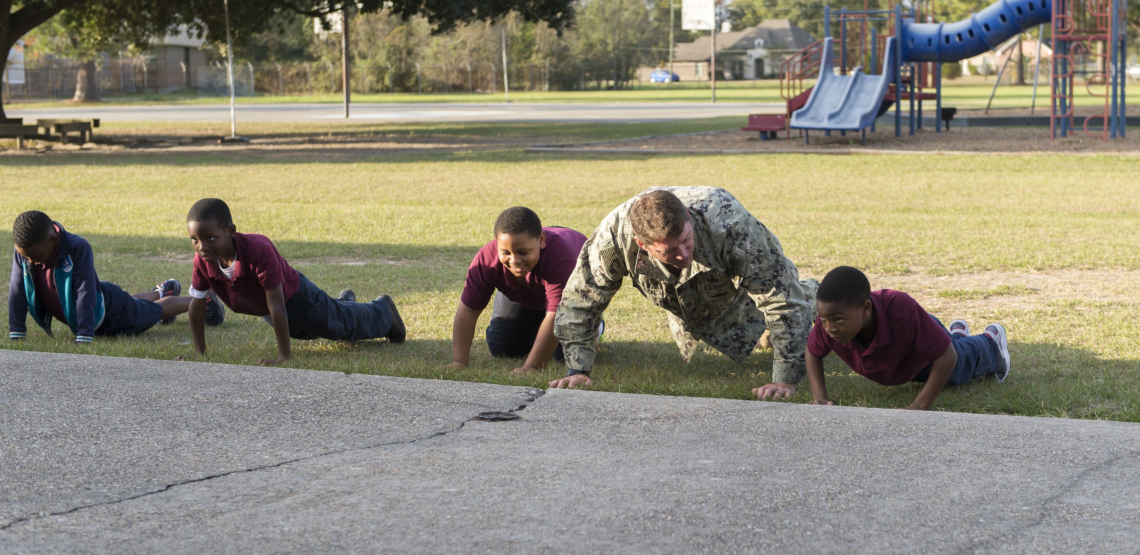 Kinder machen Liegestütze auf einer Wiese in einem Park mit Spielplatzgeräten, Häusern, einem Auto, Bäumen und einem bewölkten Himmel.