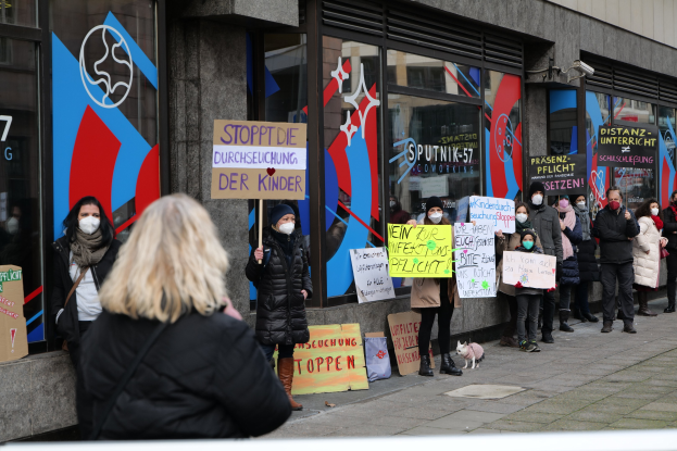Eine Gruppe von Menschen mit Masken und Schildern protestiert vor einem Gebäude mit Glaswänden und -türen, auf denen Texte und Logos zu sehen sind, sowie Kameras und einem Hund.