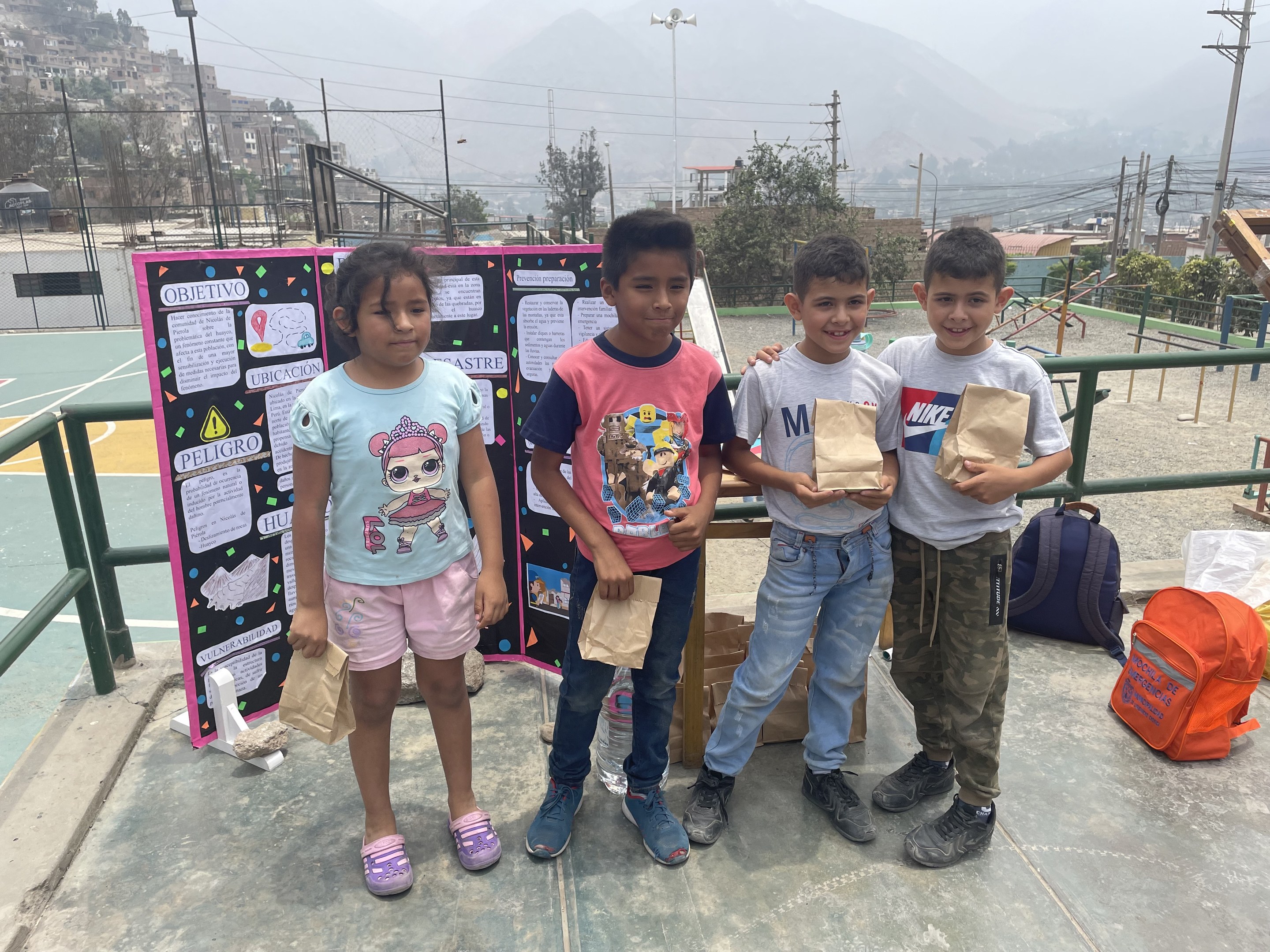 Gruppe von Kindern auf einem Basketballfeld mit Papiertüten in den Händen, Tüten und Gegenstände rechts und eine Tafel mit Text links, vor einem Hintergrund aus Zaun, Pfosten, Straßenlaternen, Bäumen, Gebäuden, Hügeln und Himmel.