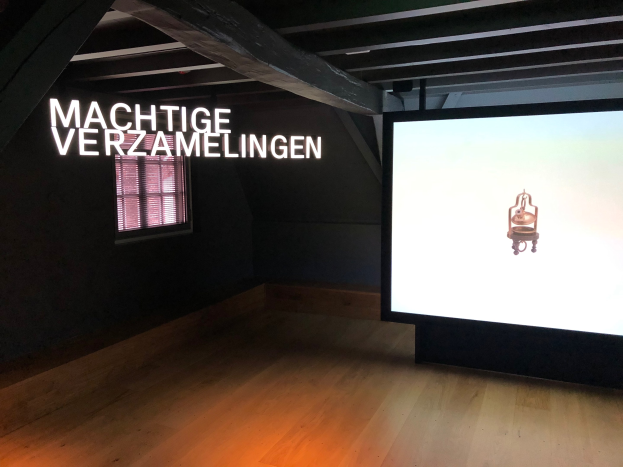 Installationsansicht der Ausstellung 'Machtige Verzamelingen' im Museum of Modern Art in Berlin, mit einem großen Bildschirm mit Text auf der rechten Seite und einem Fenster auf der linken Seite.