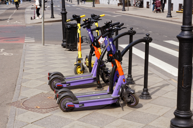 Eine Reihe von Elektro-Scootern, die an einer Stra├če mit Fü├čgängern, Radfahrern, einem Müllcontainer im Vordergrund und Geb├Ąuden, B├Ąumen und Pflanzen im Hintergrund geparkt sind.