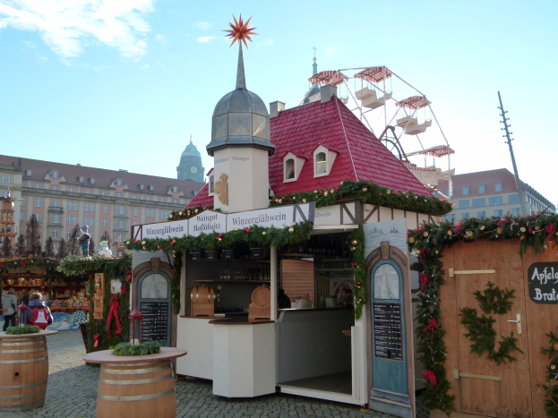 Ein kleines Gebäude mit einem roten Dach und einem Riesenrad davor, umgeben von Menschen und Festdekorationen bei einem Oktoberfest-Event in München, Deutschland.