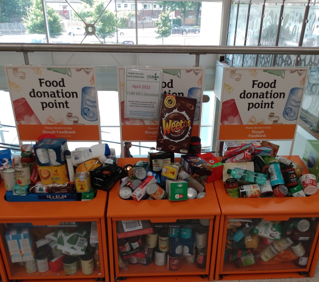 Orange Schübe mit Lebensmittelspenden, darunter Flaschen, Dosen und Kartons, mit Informationsschildern, durch Glasfenster mit Fahrzeugen, Bäumen, Gebäuden und Himmel.