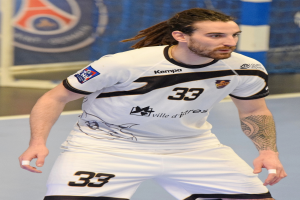 Ein Mann mit langen Haaren und Armschmuck spielt Handball in einem weißen T-Shirt und Shorts, mit einem Netz und einer Wand im Hintergrund.