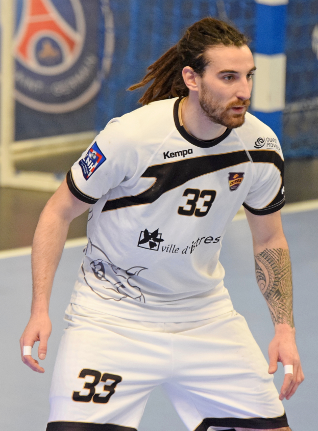 Ein Mann mit langen Haaren und Armschmuck spielt Handball in einem weißen T-Shirt und Shorts, mit einem Netz und einer Wand im Hintergrund.