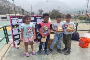 Eine Gruppe von Kindern steht auf einem Basketballfeld, jedes Kind hält eine Papiertüte in der Hand, mit Tüten und Gegenständen rechts daneben und einer Texttafel links daneben, mit Zäunen, Pfosten, Straßenlaternen, Bäumen, Gebäuden, Hügeln und dem Himmel im Hintergrund.