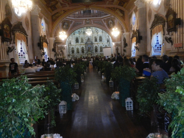 Eine Hochzeitszeremonie in einer Kirche mit Sitzplätzen und Stehplätzen, Topfpflanzen, die den Gang schmücken, und Dekorationselementen wie Skulpturen, Vorhänge, Kronleuchter und ein Bildschirm im Hintergrund.