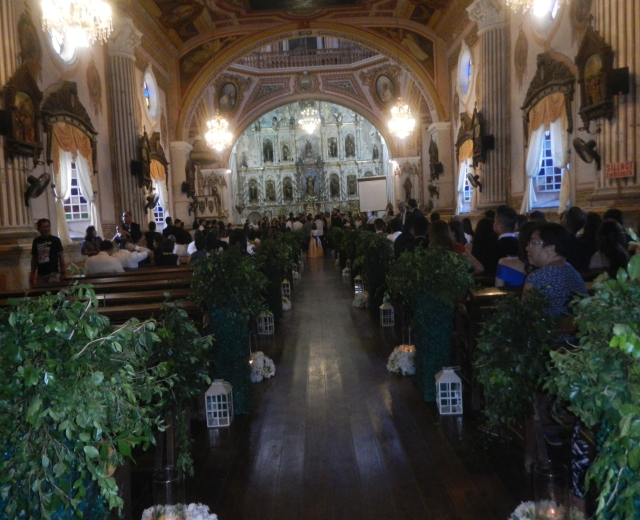 Eine Hochzeitszeremonie in einer Kirche mit Sitzplätzen und Stehplätzen, Topfpflanzen, die den Gang schmücken, und Dekorationselementen wie Skulpturen, Vorhänge, Kronleuchter und ein Bildschirm im Hintergrund.