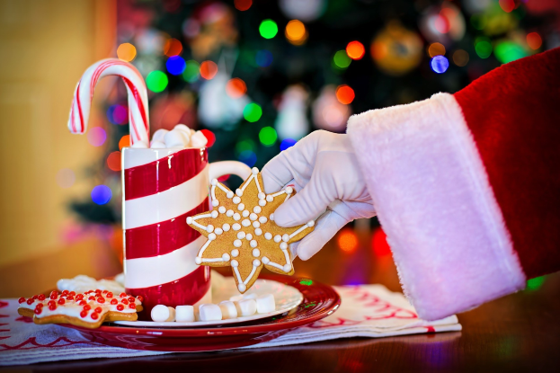 Mann in Santa Claus Mütze mit Tasse heißer Schokolade und Bonbon, mit Plätzchen und Tuch auf einem Tisch, vor einem Weihnachtsbaum und Wand.