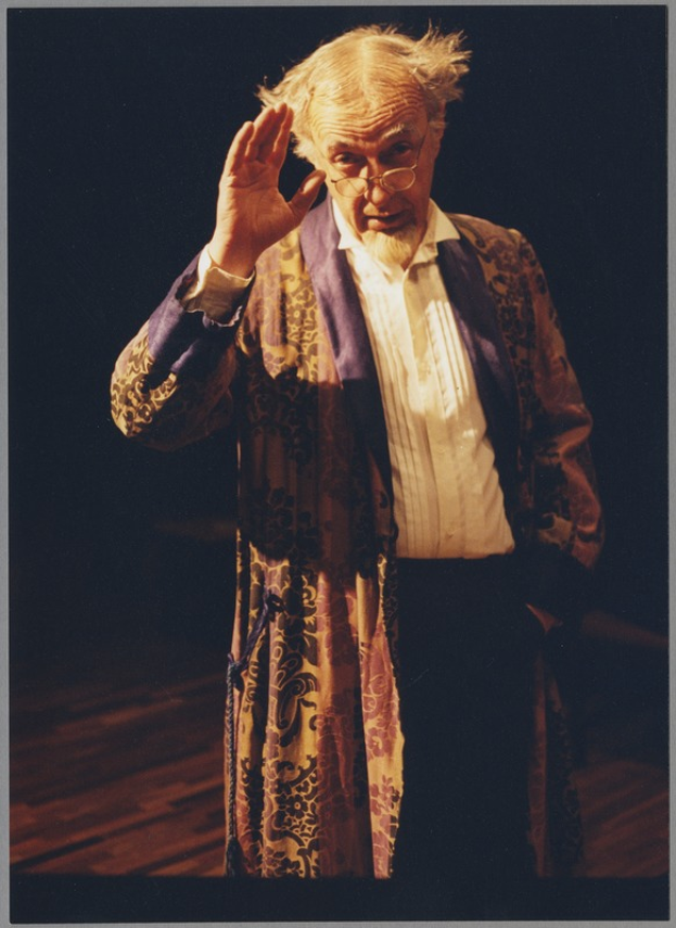 Ein Mann in einem Gewand und Brille steht auf einer Bühne mit dunklem Hintergrund, hält seine Hände zusammen und schaut ernst ins Publikum.