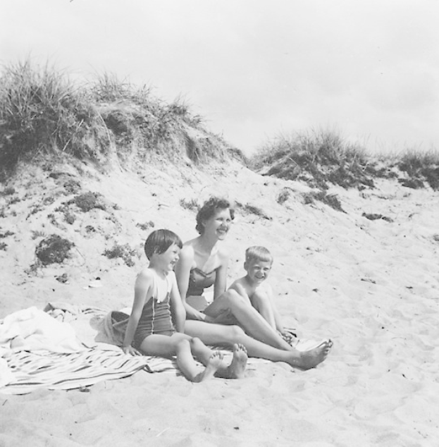 Schwarzes und weißes Foto einer Frau in einem Bikini und zwei Kindern in Badebekleidung auf einer Decke am Strand sitzend, umgeben von Sand und Gras mit Himmel darüber.