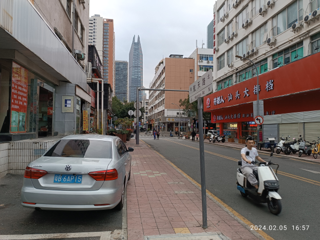 Ein Mann fährt mit einem Roller eine städtische Straße entlang, die von hohen Gebäuden gesäumt ist, mit Fußgängern auf den Gehwegen und anderen Fahrzeugen auf der Straße, unter einem klaren blauen Himmel mit Bäumen im Hintergrund.