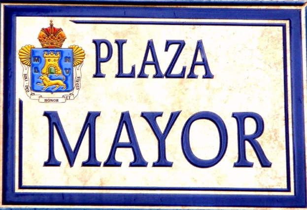 Rechteckige weiße Plakette mit schwarzem Text "Plaza Mayor" in fetter Schrift, blauem Rand und einem zentralen gekrönten Wappen-Logo, das an einem Gebäude angebracht ist.