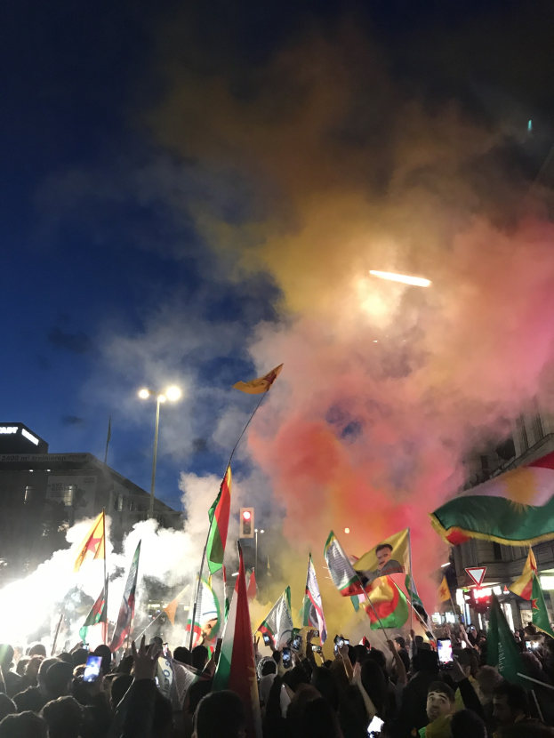 Menschenmenge vor einem Gebäude mit Fahnen schwenkend und Handys haltend unter farbigen Feuerwerkskörpern im Nachthimmel.