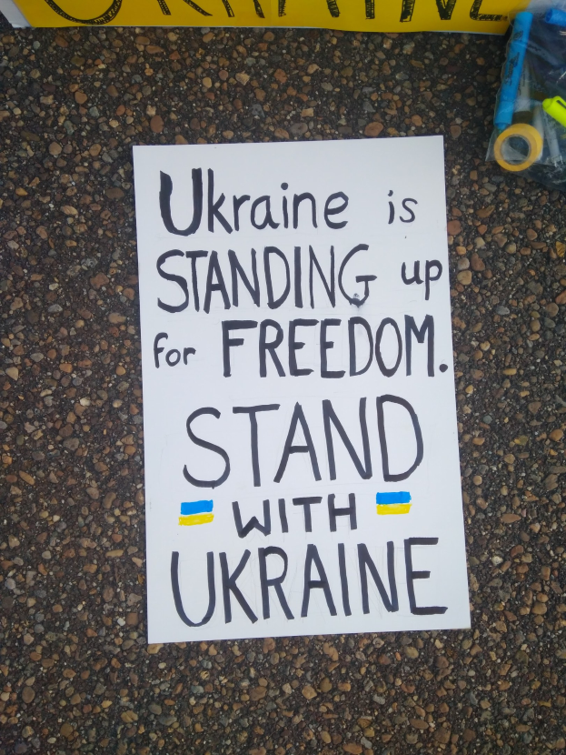 Ein weißes Blatt mit dem Text "Ukraine steht für Freiheit ein, stellt dich mit Ukraine" liegt vor einer gelben Fahne auf dem Boden. Rechts davon befindet sich eine Plastikhülle mit verschiedenen Gegenständen.