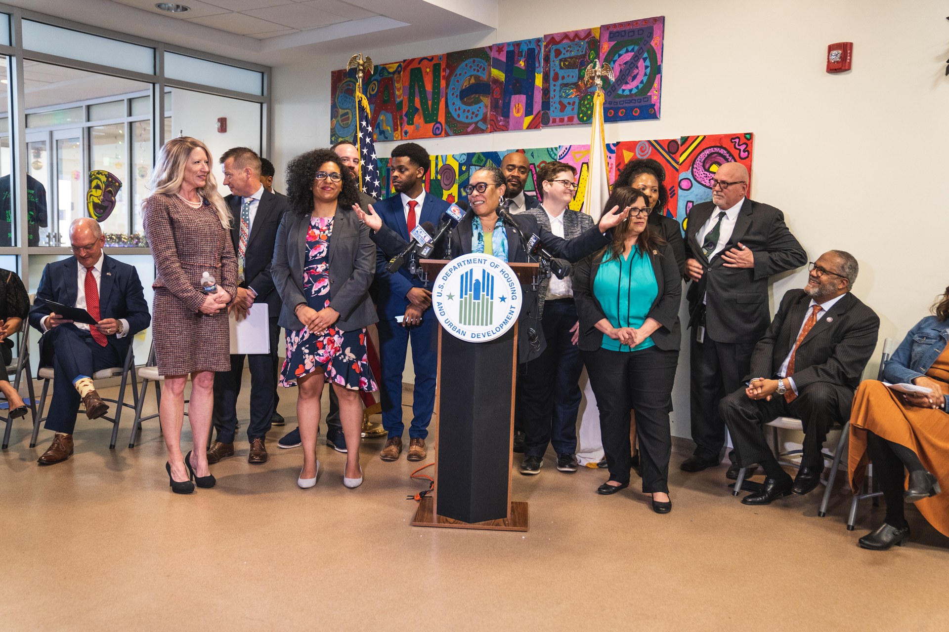 Gruppe von Menschen vor einem Rednerpult mit Mikrofon und einer Tafel mit der Aufschrift "Sancho-Stadtrat tritt dem Stadtrat bei", mit sitzenden und stehenden Teilnehmern, Flaggen, Feuerlöscher und Deckenbeleuchtung im Hintergrund.