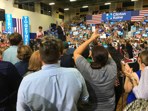 Eine große Menge jubelt vor einer Bühne, auf der ein Mann in ein Mikrofon spricht und eine Frau neben ihm steht, mit Flaggen, Transparenten und einem Lautsprecher im Hintergrund der Bühne.