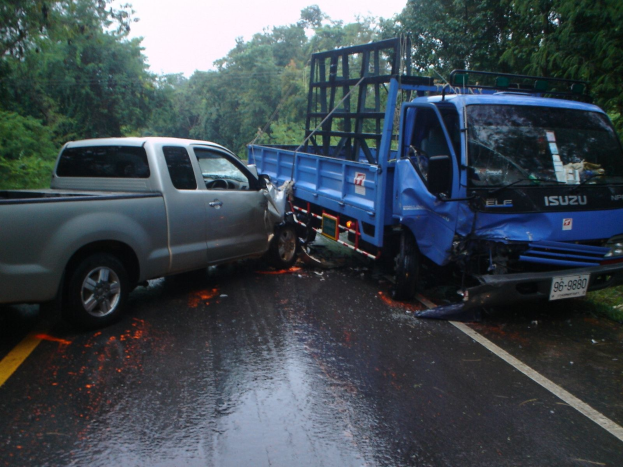 Ein demolierter Lkw mit eingedrückter Front und verbeulter Karosserie liegt auf der Seite einer Straße, umgeben von Bäumen unter einem klaren blauen Himmel.