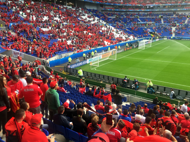 Ein volles Stadion mit Zuschauern, einige sitzen und einige stehen, die ein Fußballspiel beobachten, mit Bannern, einem Torpfosten und Deckenleuchten im Hintergrund.