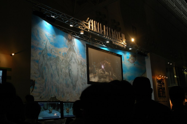 Eine Bühne mit einem Video-Display in der Mitte, vor der eine Gruppe von Menschen sitzt.