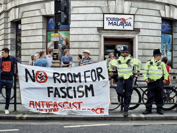 Eine Gruppe von Menschen mit einer 'Kein Platz für Faschismus'-Plakette, umgeben von zwei Polizeibeamten, Fahrrädern, einer Ampel und einem Gebäude im Hintergrund.