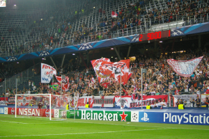 Ein Fußballspiel wird in einem hell erleuchteten Stadion mit einer großen Zuschauermenge, grünem Rasen, einem Tor mit Bannern und einem Bildschirm in der oberen linken Ecke gespielt.