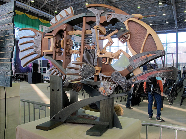 Große Metallfischskulptur drinnen ausgestellt mit Zuschauern, umgeben von Stühlen, einem Zaun, einer Fahne, einem Lautsprecher, Vorhängen und Deckenlampen.