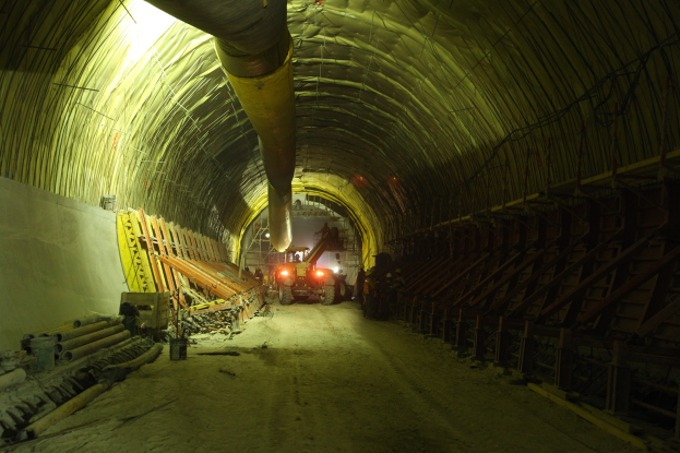 Baustelle mit einem zentralen Tunnel, Fahrzeugen, verstreuten Materialien, einer Wand auf der linken Seite, Überkopfrohren und beleuchtetem Tunnelhintergrund.
