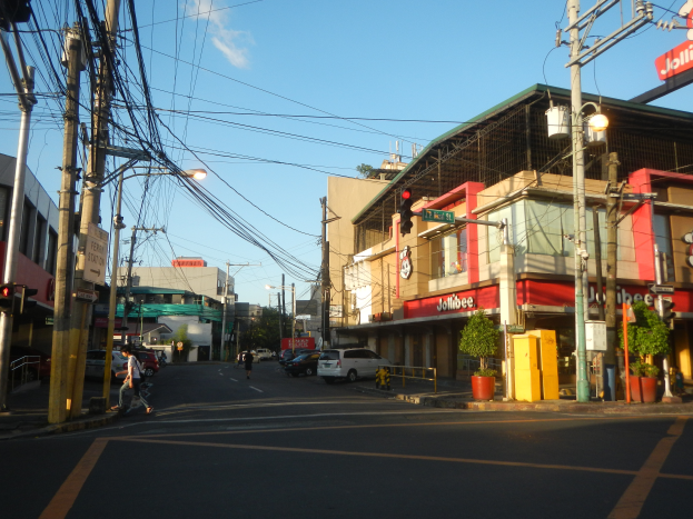 Eine Stadtstraßenszene mit Autos, Fußgängern, Versorgungsmasten, Ampeln, Gebäuden mit Schildern, Topfpflanzen und einer bewölkten Himmel.