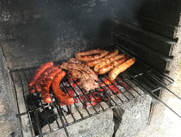 Grill mit Würstchen und Fleisch darauf, umgeben von brennender Kohle und Flammen, draußen auf Gras.