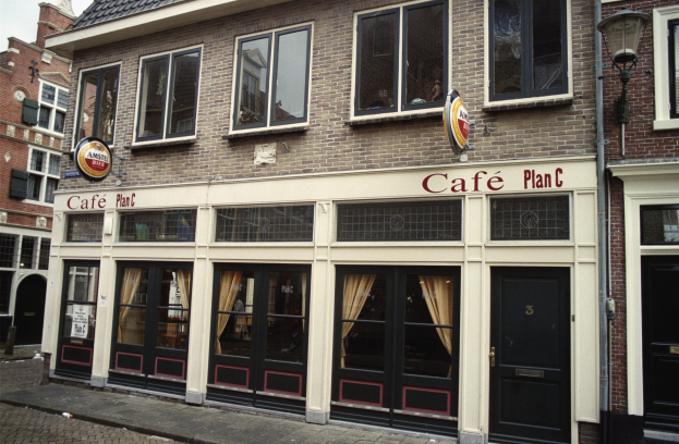 Ein Gebäude mit Fenstern und Türen, ein Schild mit der Aufschrift "Cafe Plan C in Amsterdam, Niederlande" an einem Laternenpfahl, umgeben von Bäumen unter einem bewölkten Himmel.