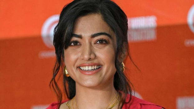 Eine Frau in einem roten Kleid mit einem eleganten Dutt und dezentem Make-up steht lächelnd da, trägt schwarze High Heels und hat ein hellgelbes Banner mit der Aufschrift 'Sonam Kapoors Schönste Looks vom Cannes Film Festival' im Hintergrund.
