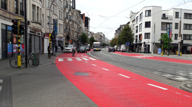 Rote Fahrradspur neben einer städtischen Straße mit fahrenden Fahrzeugen, Fußgängern auf dem Gehweg, Gebäuden mit Fenstern, Bäumen und einem klaren blauen Himmel im Hintergrund.