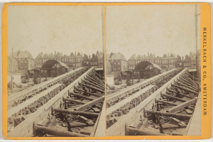 Ein altes Schwarz-Weiß-Foto einer Bahntrasse mit Gebäuden im Hintergrund, Text auf der rechten Seite lesbar "Merkelbach & Co, Co. Amsterdam" und verstreute hölzerne Gegenstände im Vordergrund.