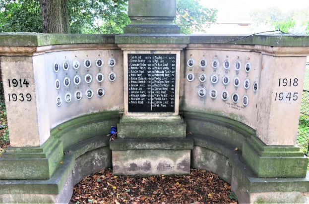 Ein Holocaust-Gedenkmonument in einem jüdischen Friedhof in Berlin, mit einer Tafel mit Text und Zahlen, umgeben von Bäumen und einem Zaun mit verstreuten trockenen Blättern.
